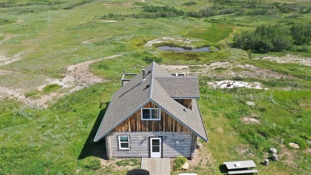 Eine Holzhütte mit Satteldach, eingebettet in eine grüne Landschaft mit einem kleinen Teich und weitläufigen Feldern.