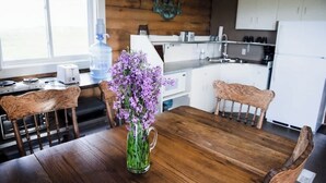 Ein Holztisch mit Stühlen, eine Vase mit lila Blumen, ein Wasserspender und eine Küche im Hintergrund.