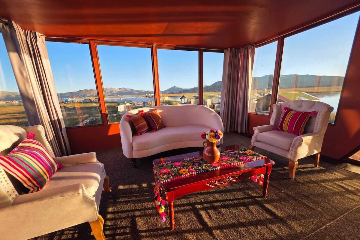A room with a large window, two white sofas, a wooden coffee table with a floral centerpiece, and a view of a landscape.