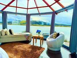 A room with a view of a lake and hills, a sofa, a chair, a small table with a vase, and a rug.