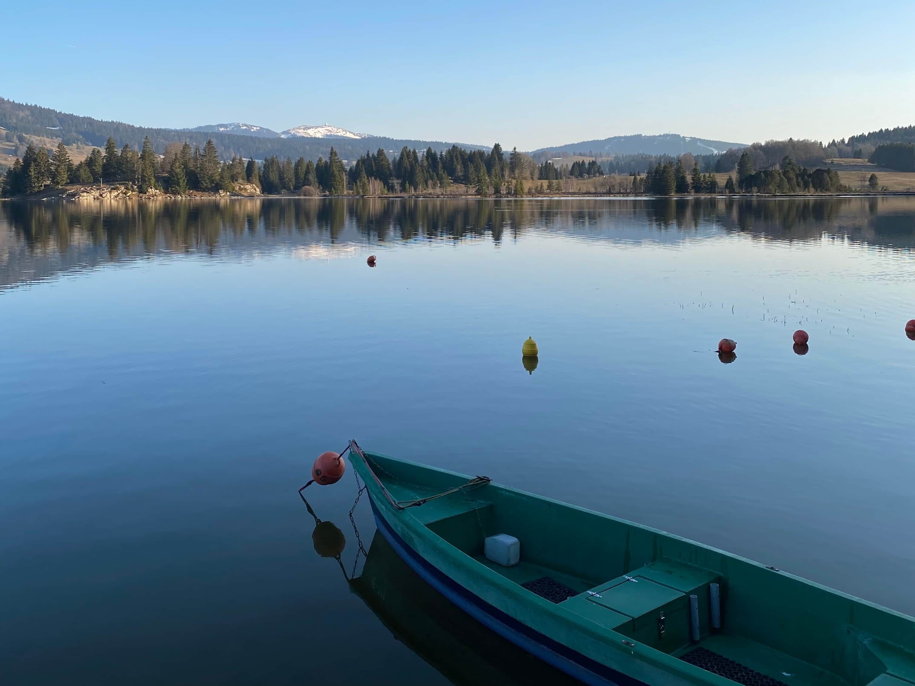 A calm lake with a green boat docked at the edge, surrounded by trees and distant mountains.