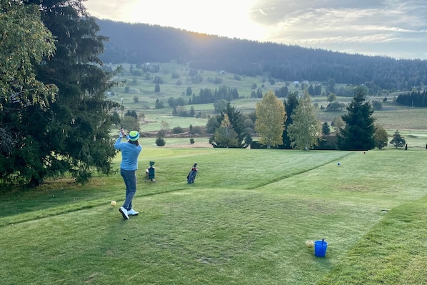 A golf course with a person preparing to hit a ball, a blue bucket, and a golf cart.