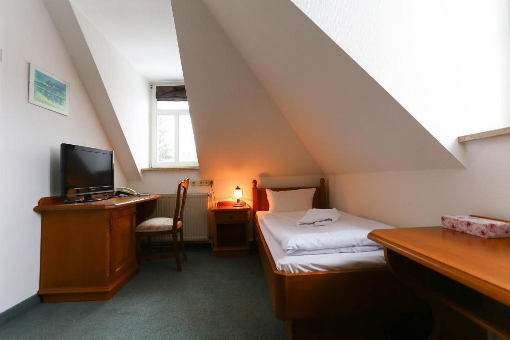 Una piccola stanza triangolare con un letto, una scrivania con televisore, una sedia e un comodino con lampada.