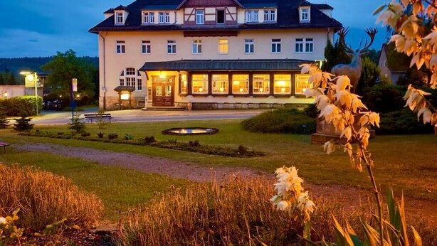 Ein großes, mehrstöckiges Gebäude mit einer symmetrischen Fassade, einem zentralen Eingang und einem gepflegten Garten mit Teich.