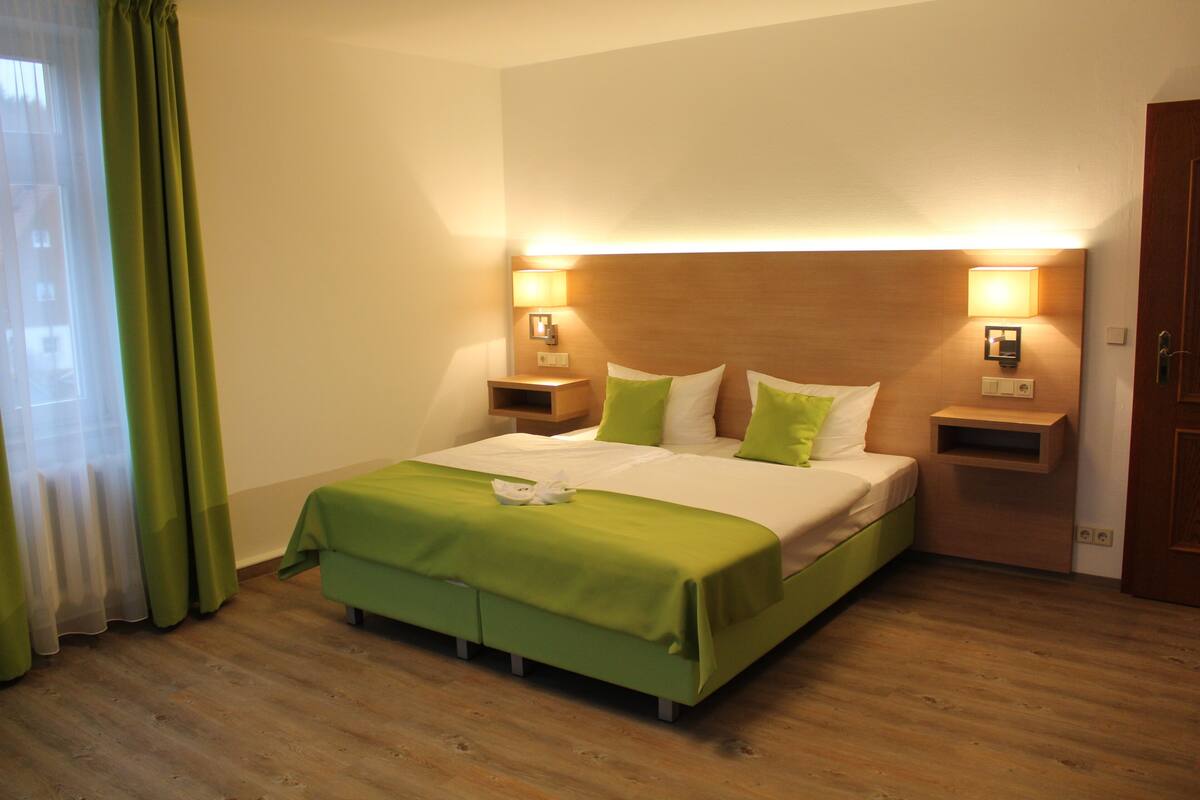 A hotel room with a bed, green curtains, wooden flooring, and bedside lamps.