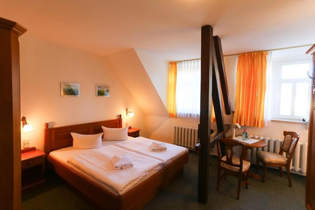 Una habitación de hotel con una cama grande, una mesita de noche, una silla, una pequeña mesa y un ventanal con cortinas.
