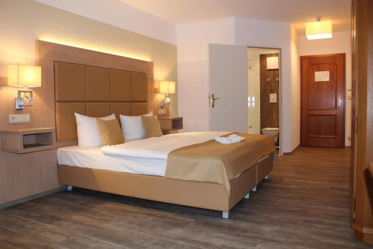 A hotel room with a large bed, bedside lamps, a wooden headboard, a wooden door, and a wooden floor.