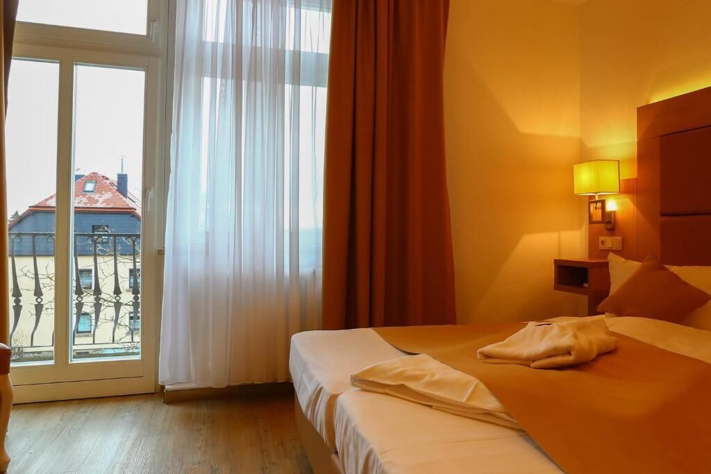 A hotel room with a bed, a nightstand, a lamp, and a view of buildings through a window.