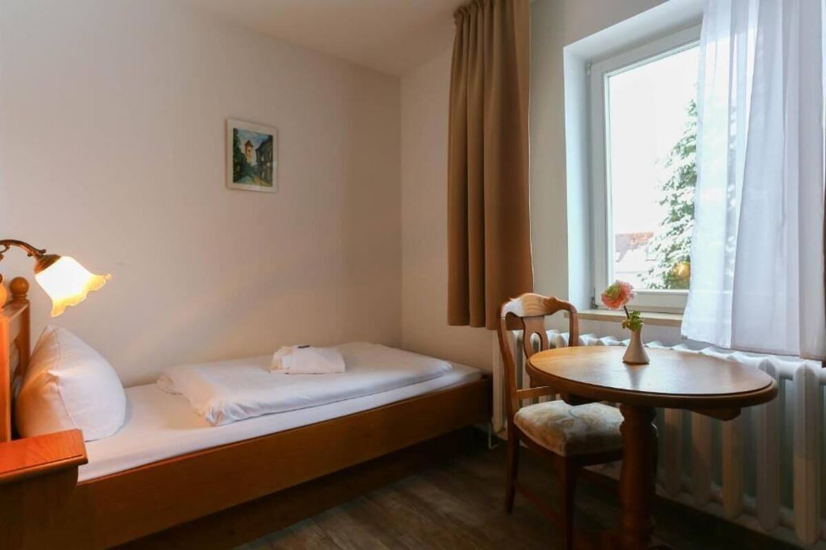 A single bed with a wooden headboard, a bedside table with a lamp, a small table with a vase of flowers, and a window with curtains.
