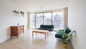 Un salon spacieux avec une table basse en bois, un canapé vert, un meuble en bois et une grande fenêtre donnant sur la ville.