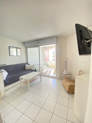 Ein Wohnzimmer mit einer Couch, einem kleinen Tisch, einem an der Wand montierten Fernseher und einer Glasschiebetür, die zu einem Außenbereich führt.