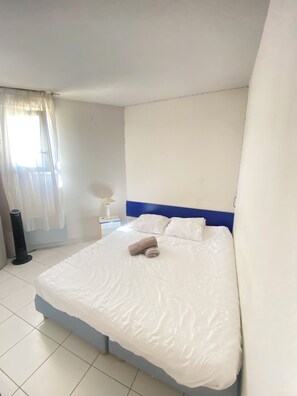 Ein Hotelzimmer mit einem großen Bett, einem Nachttisch mit Lampe, einem Fenster mit durchsichtigen Vorhängen und einem Standheizkörper.
