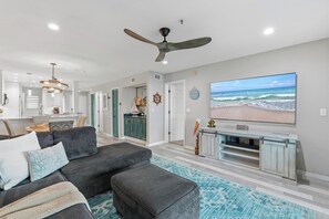 A modern living room with a sectional sofa, ottomans, a flat-screen TV, and a ceiling fan. - 🚲 Bouchelle Island 202 | Intracoastal Views | Screened Porch & Beach Cruisers (New Smyrna Beach)
