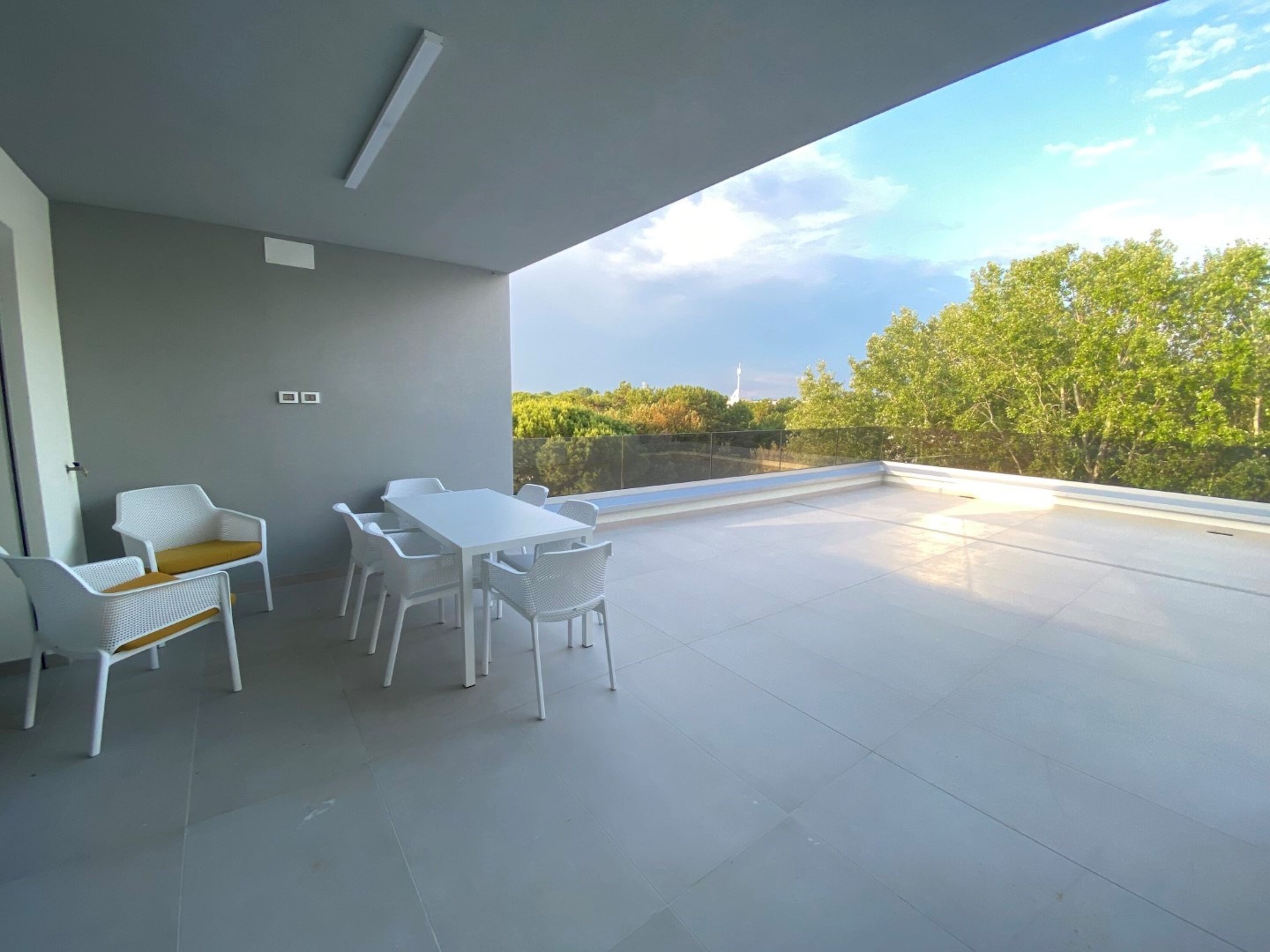 Un balcon avec une table et des chaises blanches, une vue sur des arbres et un bâtiment au loin.