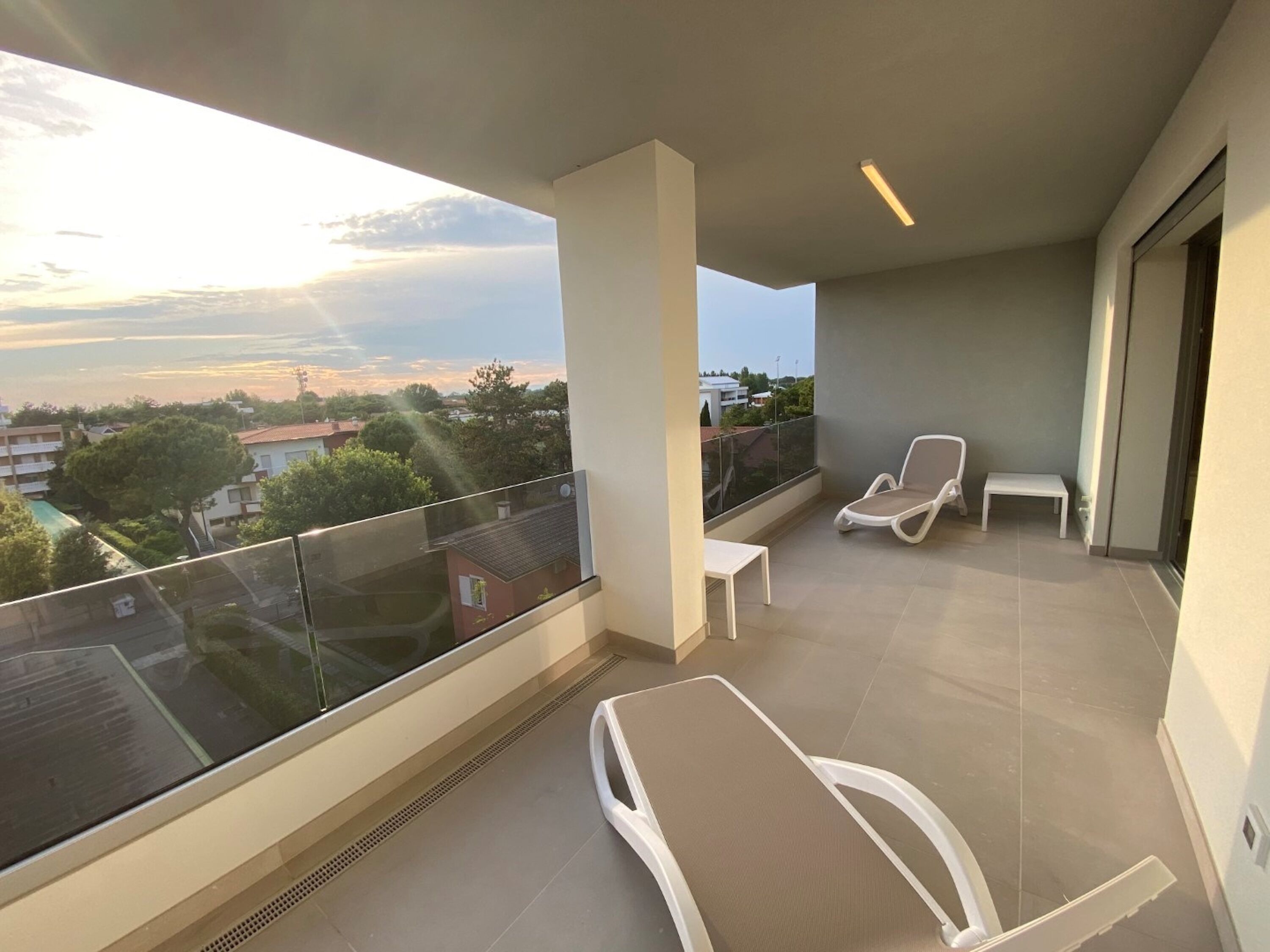 A balcony with two white lounge chairs, a small table, and a view of a residential area with trees and buildings.