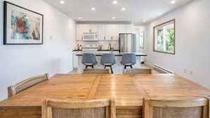 A modern kitchen with white cabinets, a stainless steel refrigerator, and a dining area with a wooden table and wicker chairs.