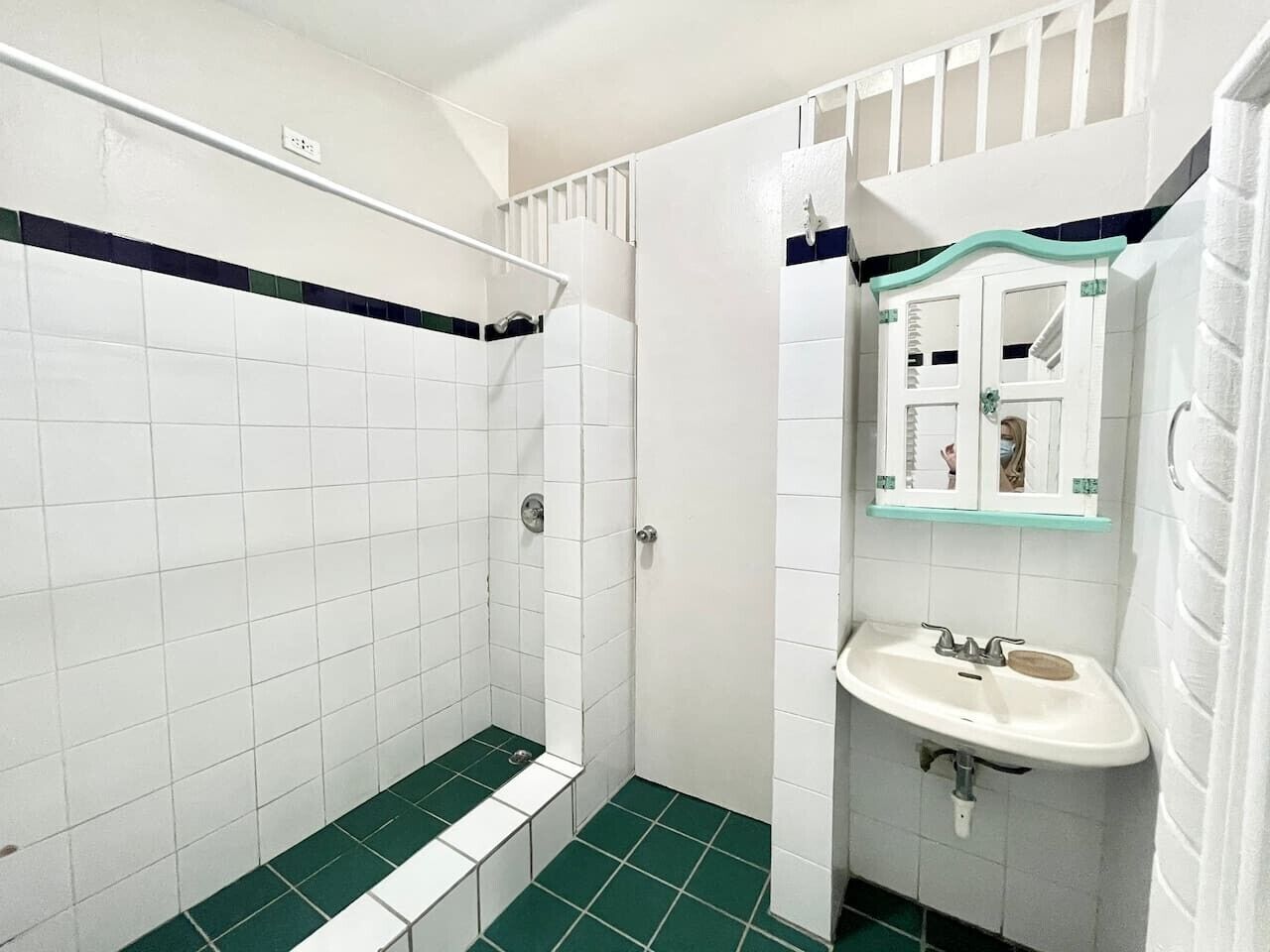 A bathroom with white tiles, a green-tiled shower floor, a white sink, and a mirror with a glass shelf above it.
