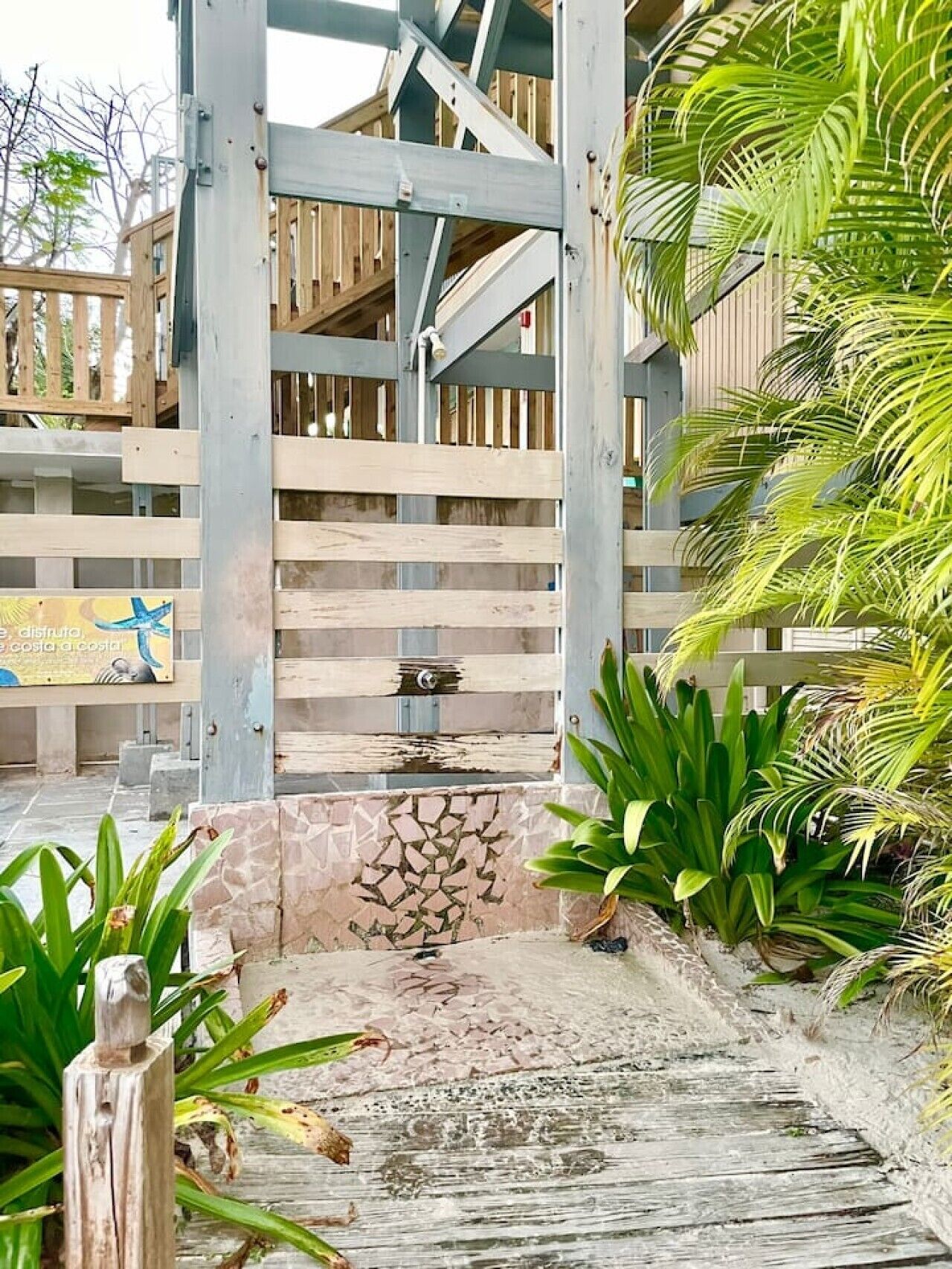 A wooden gate leading to a building with a stone wall and tropical plants.