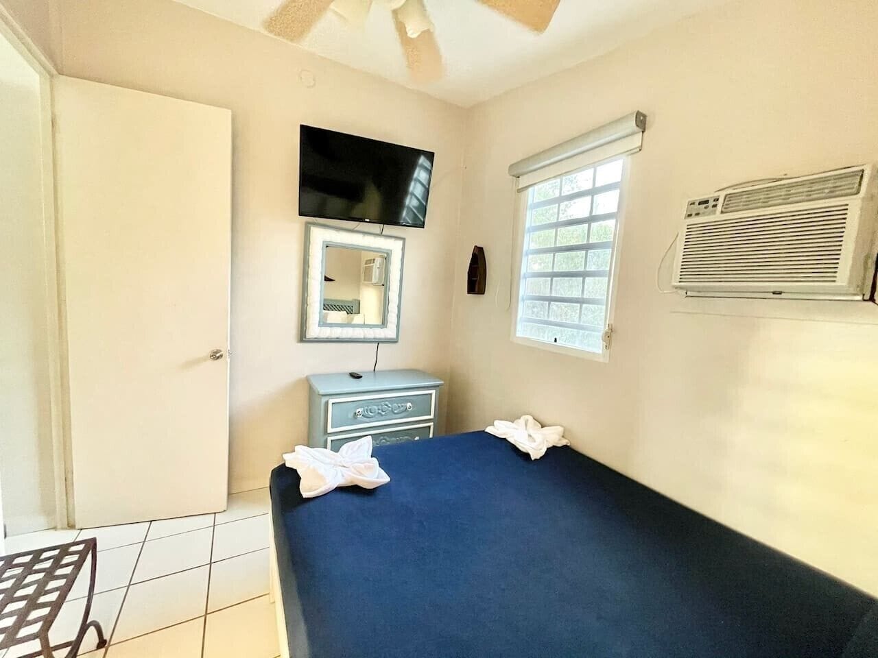 A room with a bed, a television, a window with blinds, a mirror, and a wall-mounted air conditioner.