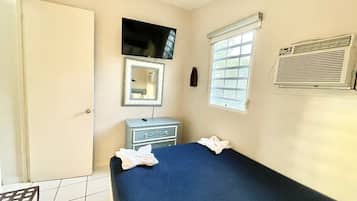 A room with a bed, a television, a window with blinds, a mirror, and a wall-mounted air conditioner.
