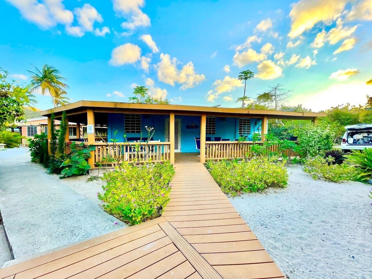 A beachfront hotel with a wooden deck leading to the entrance, surrounded by lush greenery and palm trees.