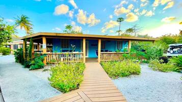 A beachfront hotel with a wooden deck leading to the entrance, surrounded by lush greenery and palm trees.