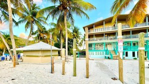 Ein Hotel direkt am Strand mit einer zweistöckigen Struktur, einer überdachten Sitzgelegenheit und Palmen.