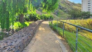 Un sendero con un muro de piedra a un lado y una barandilla metálica al otro, que conduce a una zona cubierta de césped con árboles.