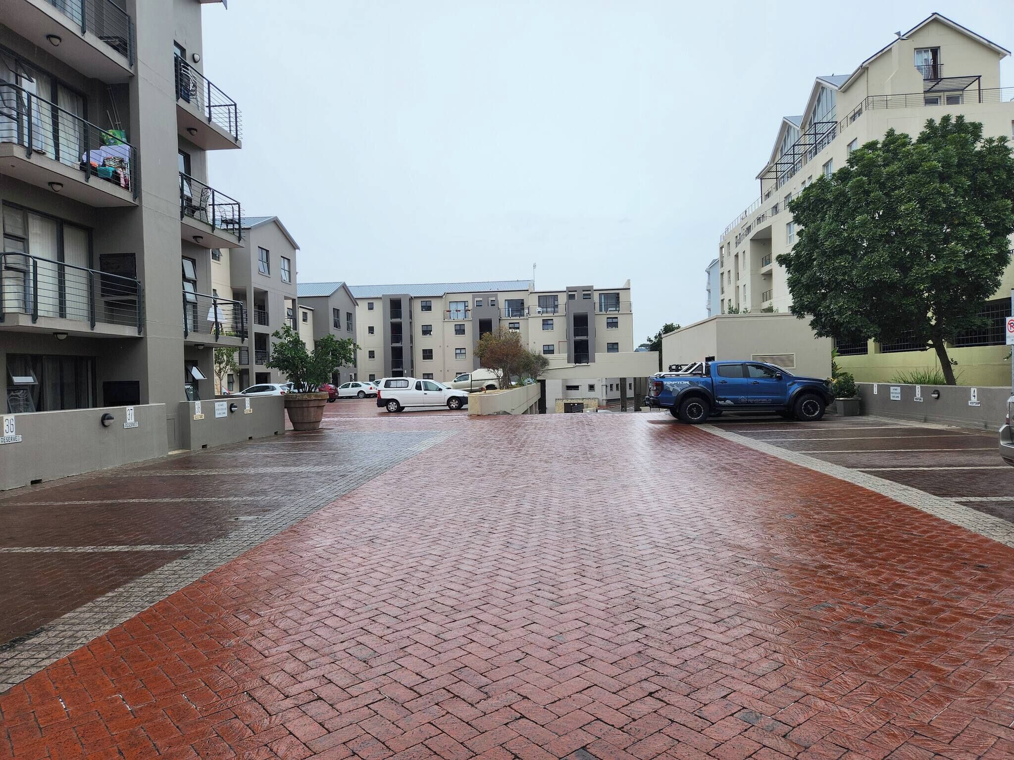 A multi-story residential building with a brick-paved courtyard, parked cars, and a blue vehicle.