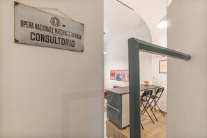Ein Schild mit der Aufschrift 'OPERA NAZIONALE MATERNITA INFANZIA CONSULTORIO' in einem Flur mit moderner Innenausstattung, einem Essbereich mit Tisch und Stühlen sowie einer kleinen Küche.