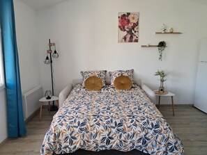 Ein Schlafzimmer mit einem blumigen Bettüberwurf, einem Nachttisch mit einer Pflanze und einem an der Wand befestigten Regal mit dekorativen Gegenständen.