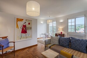 Ein Wohnzimmer mit einer Couch, einem Holztisch, einem Gemälde, einem Essbereich mit Tisch und Stühlen sowie einem Fenster mit Aussicht.