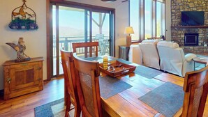 A spacious living area with a wooden dining table, comfortable seating, a stone fireplace, and large windows offering a view. - Crest 200C • Panoramic Views and Huge Shower (Sugar Mountain)