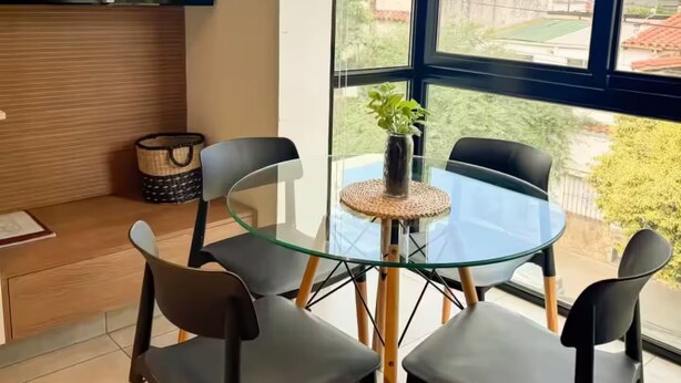 Ein modernes Zimmer mit großem Fenster, einem Flachbildfernseher, einem Glastisch mit Stühlen und einer kleinen Topfpflanze.