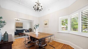 Un coin repas avec une table et des chaises en bois, un lustre et une vue dégagée sur l’extérieur grâce à des volets blancs.