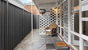 Un patio couvert avec un sol en pierre, une chaise en osier, un mur décoratif et une clôture en métal.