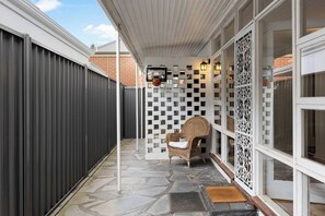 Un porche cubierto con piso de piedra, una silla de mimbre, un muro decorativo y una cerca de metal.