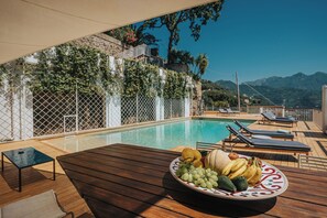 A poolside area with a wooden table, a fruit bowl, lounge chairs, and a view of mountains. - Villa Allure by My Villa Collection (Ravello)