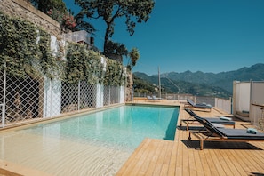 A pool with lounge chairs, a wooden deck, and a stone wall with greenery. - Villa Allure by My Villa Collection (Ravello)