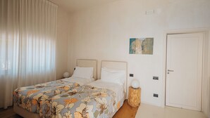 Ein Schlafzimmer mit zwei Betten, einem großen Fenster mit Vorhängen, einer Tür und einem kleinen Wandbild.