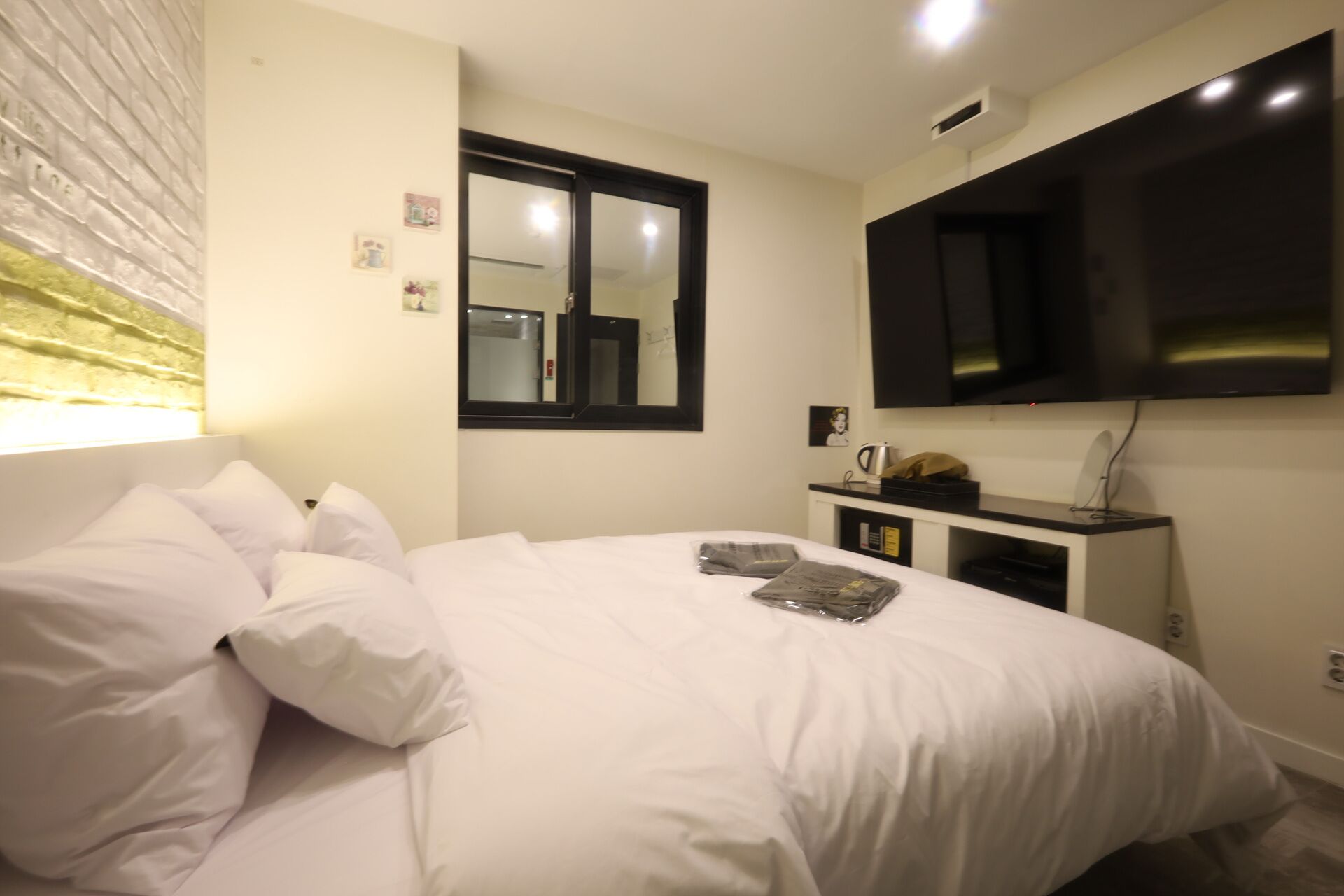 A bedroom with a bed, a TV mounted on the wall, a small bedside table with a kettle, and a window with a mirror.