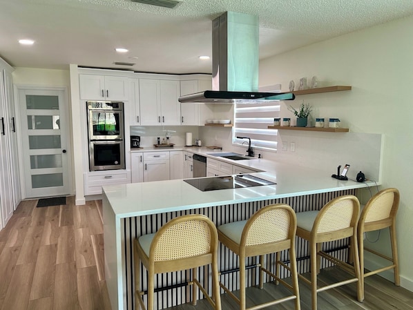 A modern kitchen with white cabinetry, a central island with bar stools, a built-in oven, and a stainless steel range hood.