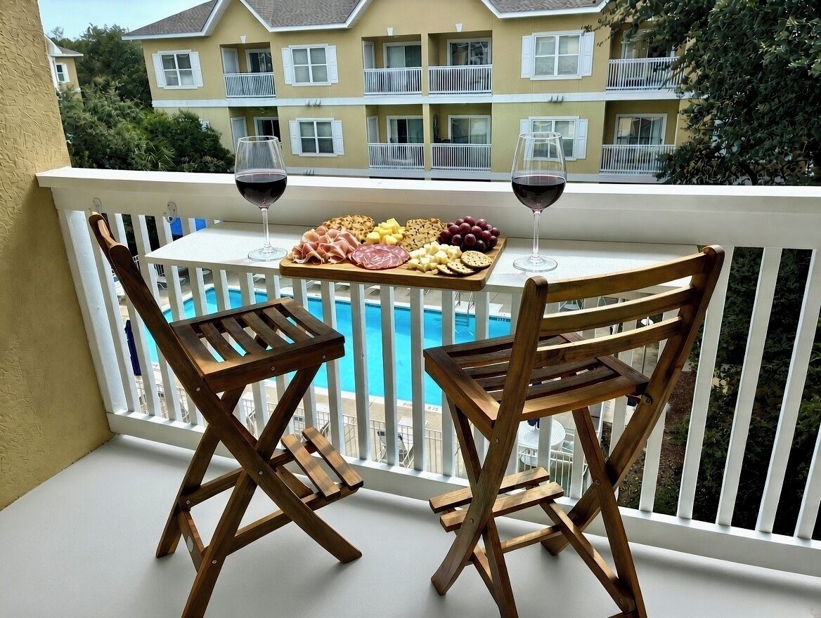 Un balcón con vista a la piscina, sillas de madera, una mesa con aperitivos y copas de vino.