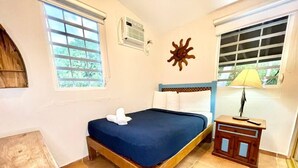 A single bed with a blue mattress, a wooden headboard, a bedside table with a lamp, and a window with blinds. - Villa 14 Flamenco Beachfront Villas (Flamenco)