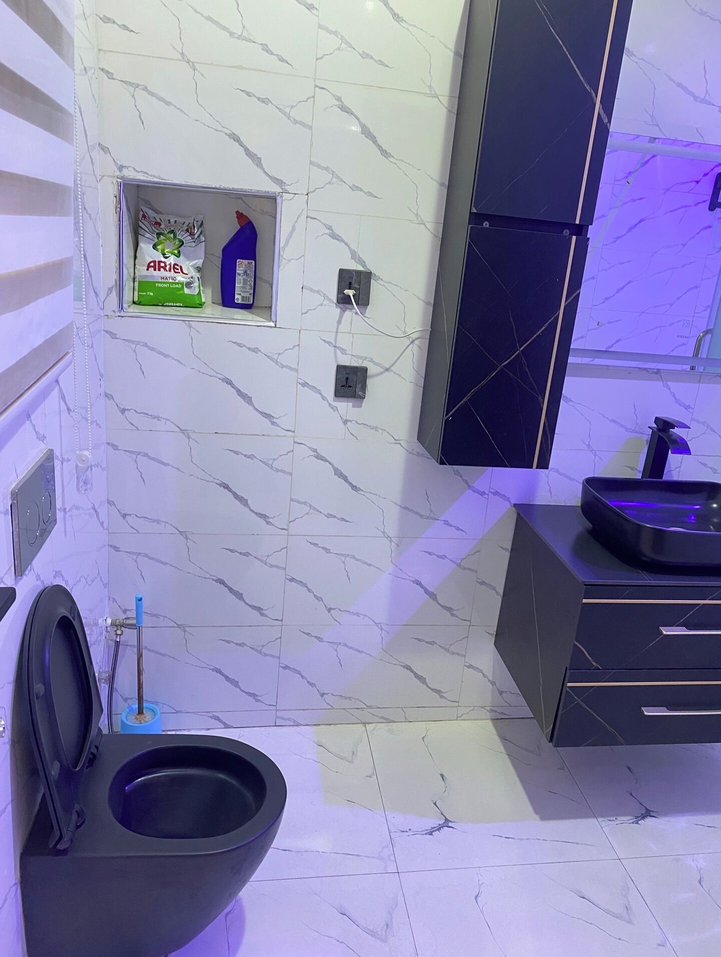 A bathroom with a black toilet, marble walls, a small shelf with toiletries, and a black cabinet.