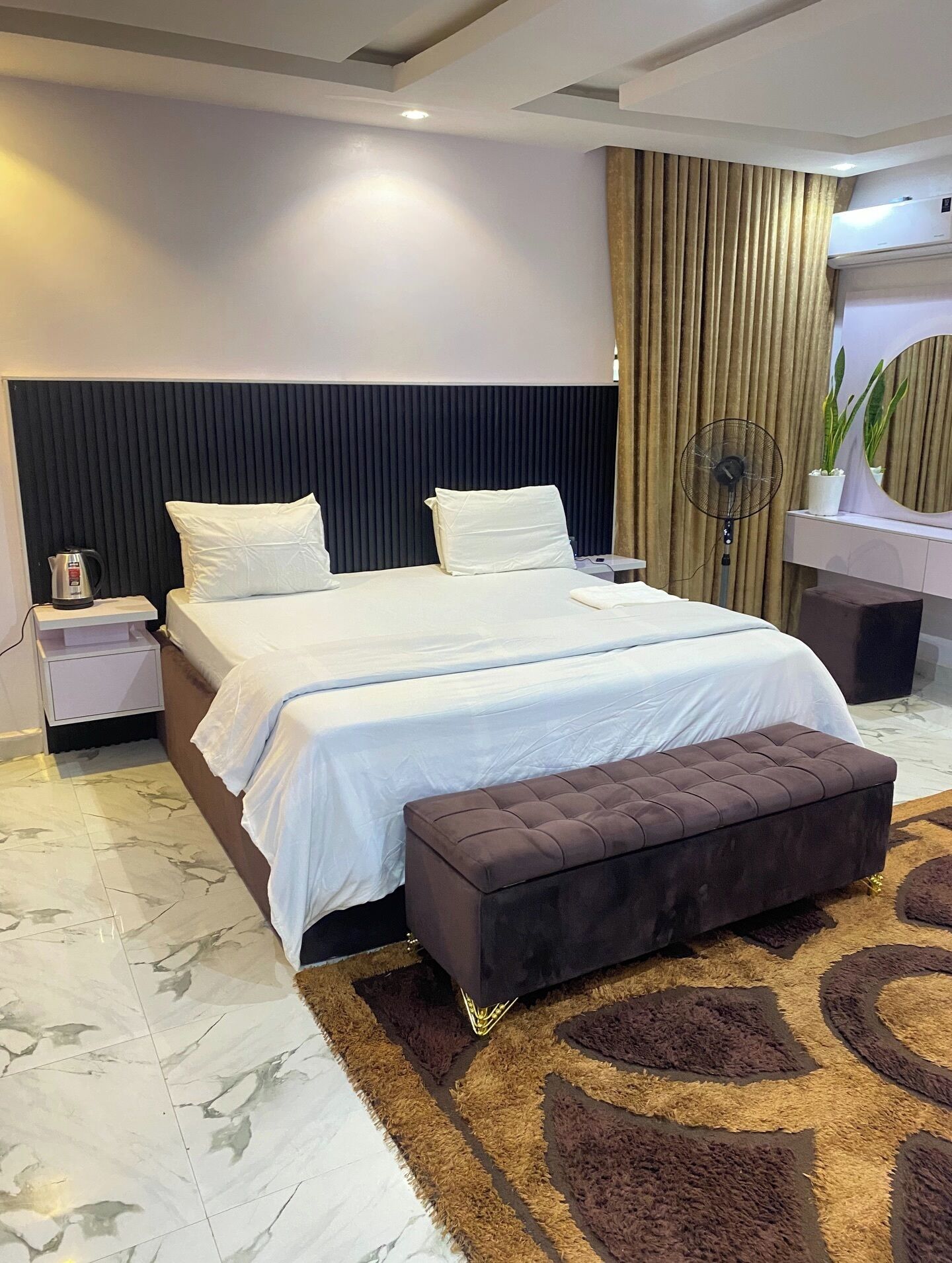 Een moderne hotelkamer met een groot bed, een nachtkastje met waterkoker, een staande ventilator en een decoratief vloerkleed.