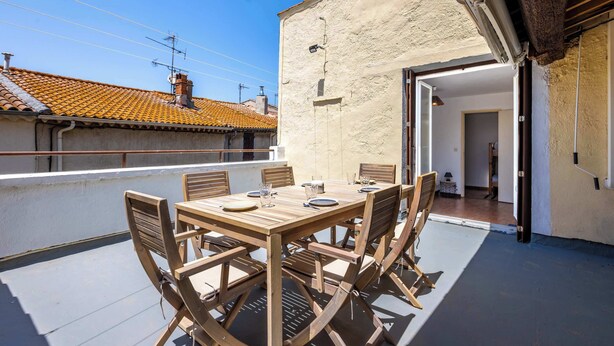Et træbord med stole på en tagterrasse med udsigt til bygninger og en klar himmel.