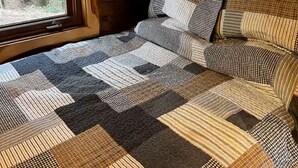 Interior de una cabaña de madera con una cama cubierta por un edredón a cuadros, ventanas amplias y vistas a un bosque.