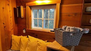 Interior de una cabaña de madera con un banco, cojines amarillos, una cesta y una ventana con vistas a los árboles.