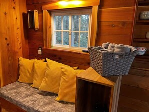 A wooden cabin interior with a bench, yellow cushions, a basket, and a window with a view of trees.